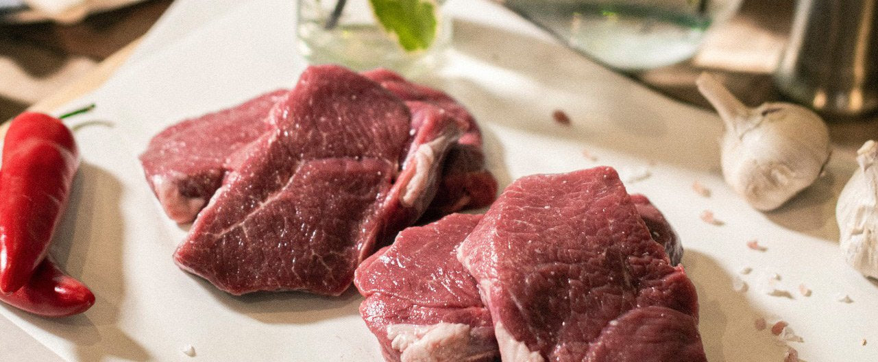 A close up of four Goodwood organic grass-fed lamb steaks on a chopping board, with red peppers and garlic cloves, at the Goodwood Farm Shop.