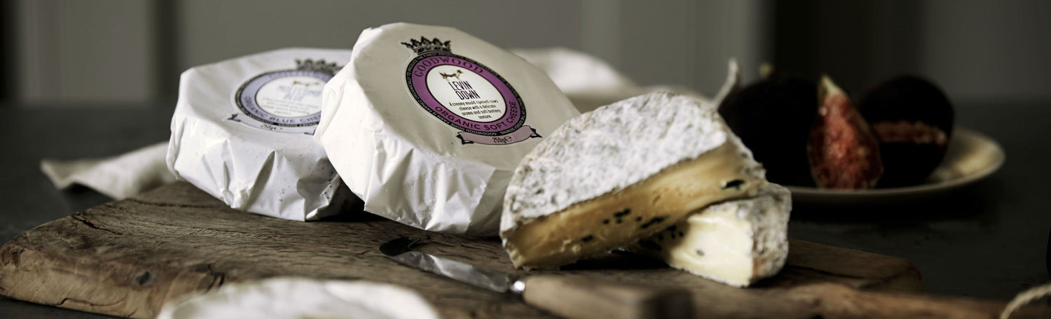 A cheese board containing Goodwood organic handmade cheeses, both packaged and sliced, including Levin Down and Molecomb Blue Goodwoof Farm Shop cheese varieties.