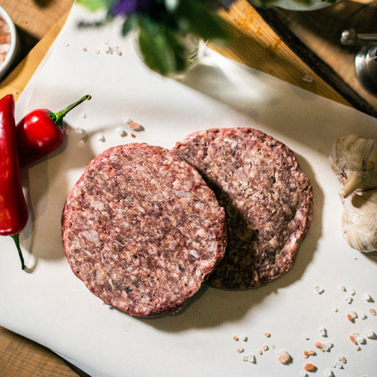 Two organic grass-fed beef burgers displayed on a chopping board, at the Goodwood Farm Shop, with red peppers and garlic cloves.
