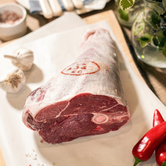 A roasting joint of organic grass-fed leg of lamb on the bone placed on a chopping board, at the Goodwood Farm Shop, with red peppers and garlic cloves.