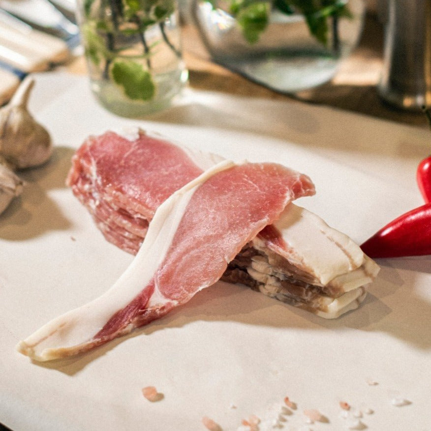 Organic West Sussex grass-fed pork back bacon rashers, shown on a chopping board with red peppers and garlic cloves.