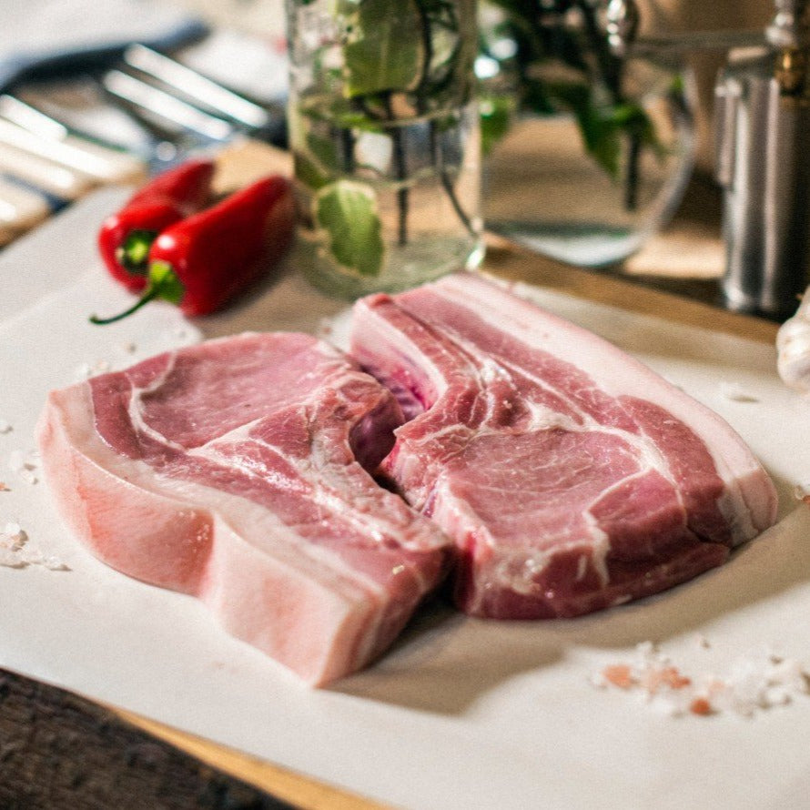 Two organic grass-fed pork loin chops displayed on a chopping board, with red peppers and garlic cloves, at the Goodwood Farm Shop.